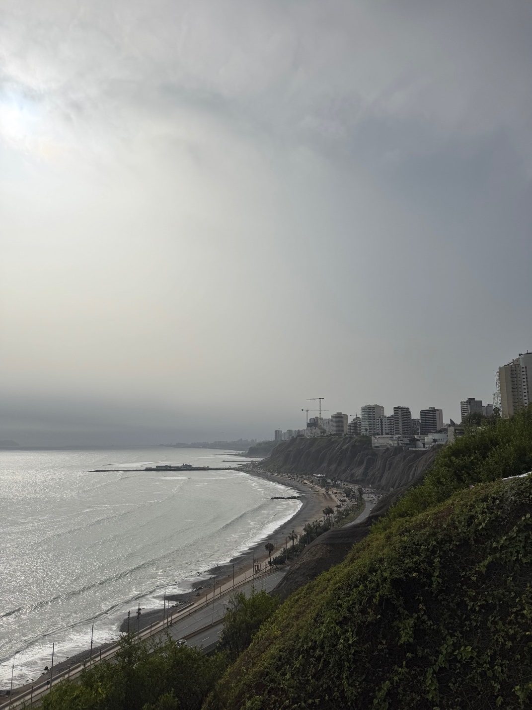 Wide view of Lima's coastline on a hazy day: a curving gray beach, coastal highway, tall eroded cliffs, and high-rise buildings with a construction crane