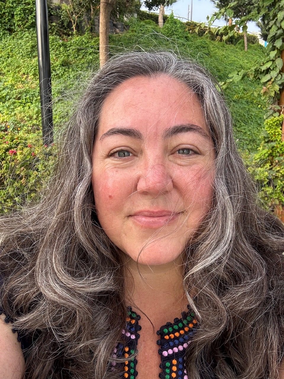 Abby Kojola smiling at the camera, long wavy gray-and-brown hair, wearing a dark embroidered top, with green hillside plantings behind her