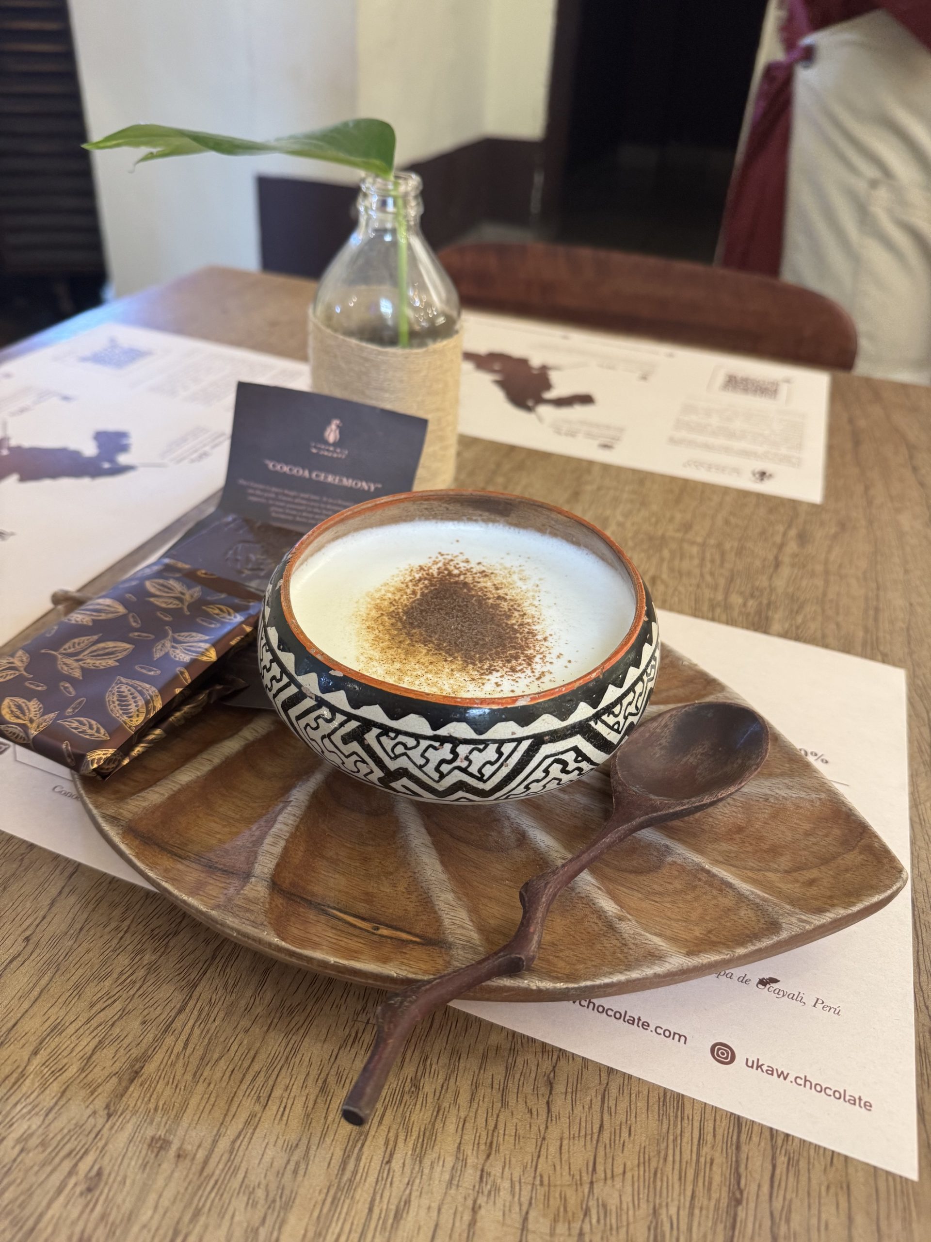 Bowl of steaming milk dusted with cinnamon in a black-and-white patterned ceramic cup, on a leaf-shaped wooden plate with a branch-like wooden spoon and a wrapped cacao bar beside it