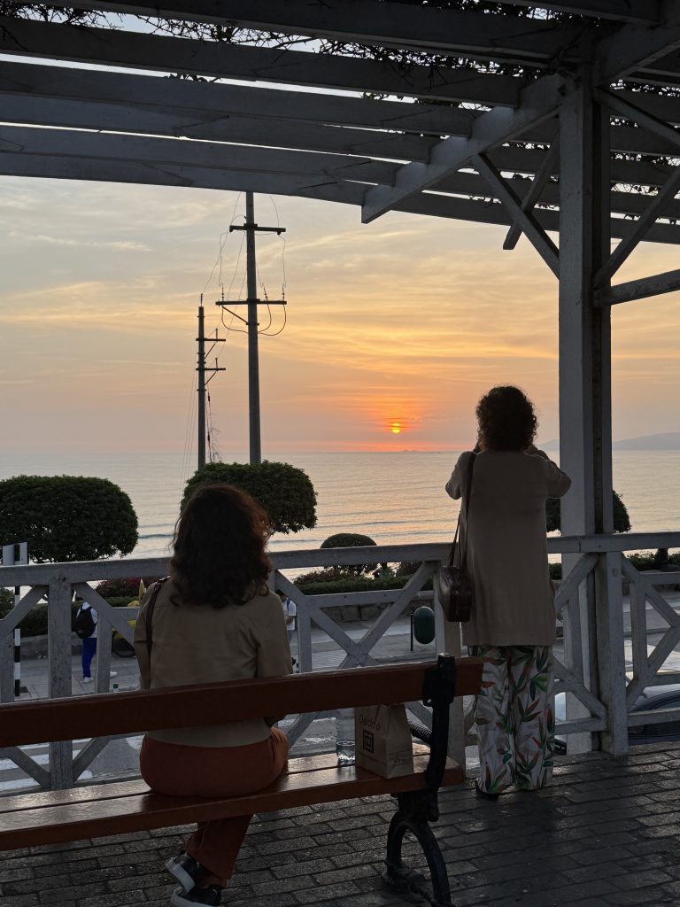 Sunset over the sea viewed from a seaside lookout; two people sit on a wooden bench under a white lattice roof, facing the water, with a bag and bottle on the bench nearby.